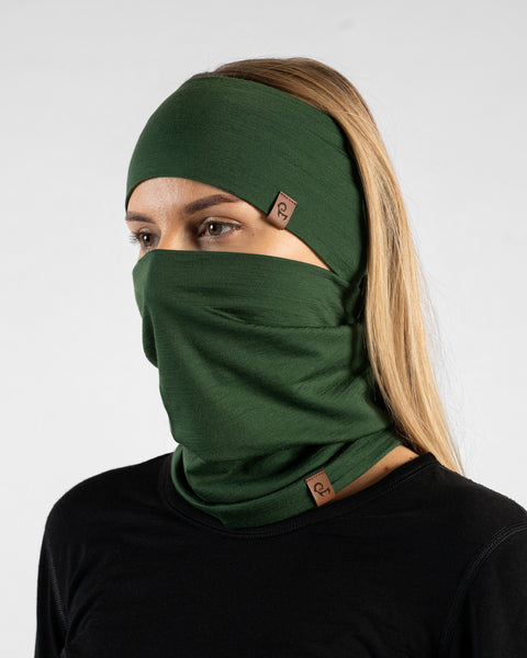 A studio portrait of a blonde woman angled slightly away from the camera, looking to the left against a plain light grey background. She is wearing a black long-sleeved shirt and a matching dark green set consisting of a wide headband and a neck gaiter pulled up over her nose and mouth. Both accessories feature small brown logo tags.