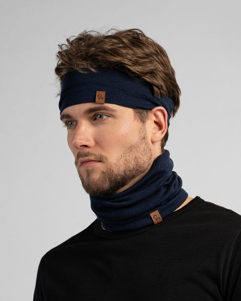 A studio portrait of a man looking to the left against a light grey background. He is wearing a black shirt, a navy blue neck gaiter, and a matching navy blue headband.