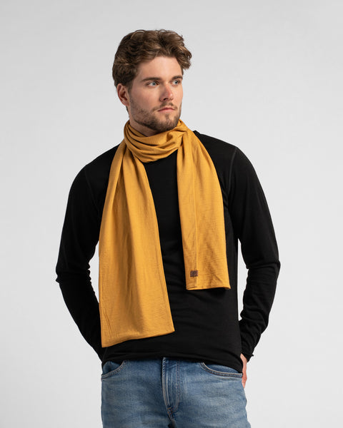 A studio portrait of a man looking off to his left, with his hands in his blue jeans pockets. He is wearing a fitted black long-sleeved shirt and a mustard yellow scarf wrapped once around his neck. The scarf's ends drape down his chest, and a small, square brown leather logo patch is visible on the bottom edge of one side. The background is a solid light grey.