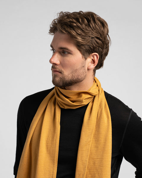 A studio portrait of a man looking off to the left side of the frame. He is wearing a black long-sleeved shirt and a mustard yellow scarf wrapped loosely around his neck against a plain light grey background.