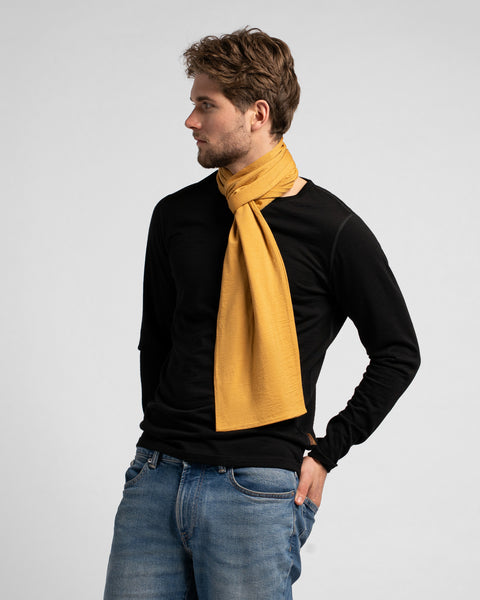 A man stands looking off to the left, with his left hand resting in his front jeans pocket. He is wearing a black long-sleeved shirt, blue jeans, and a mustard yellow scarf tied in a simple knot around his neck.