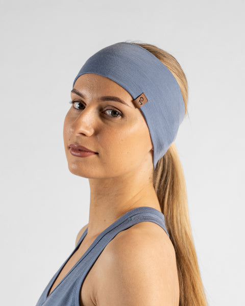 A studio portrait of a blonde woman looking directly at the camera with her body angled slightly away. She is wearing a dusty blue headband over her pulled-back hair, featuring a small brown leather logo patch. She is wearing a matching dusty blue sleeveless top against a plain light grey background.