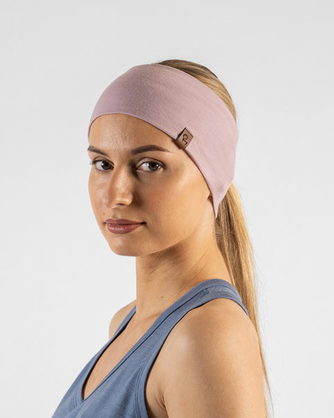 A studio portrait of a blonde woman looking directly at the camera. She is wearing a dusty pink headband over her hair, which is pulled back, featuring a small brown leather logo patch. She is wearing a dusty blue sleeveless top against a plain light grey background.