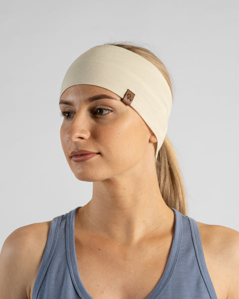 A studio portrait of a blonde woman looking off to the left side of the frame. She is wearing a light cream, natural-colored headband over her pulled-back hair, featuring a small brown leather logo patch. She is wearing a dusty blue sleeveless top against a plain light grey background.