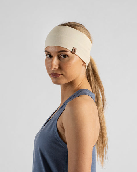 A studio portrait of a blonde woman shown in a three-quarter profile looking back over her shoulder at the camera. She is wearing a dusty blue sleeveless top and a natural off-white headband over her ponytail.