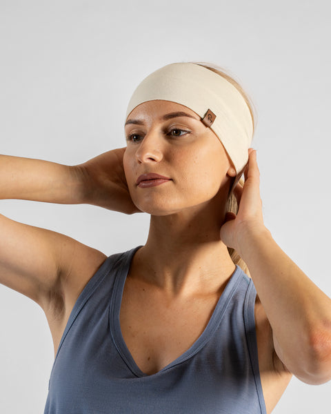 A studio portrait of a blonde woman looking off to the left against a plain light grey background. She wears a dusty blue sleeveless top and a natural off-white headband, with both hands reaching behind her head to adjust the fabric.