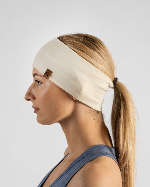 A side profile studio portrait of a blonde woman facing left against a plain light grey background. She wears a dusty blue sleeveless top and a natural off-white headband over a ponytail, with a small brown logo tag visible on the side.