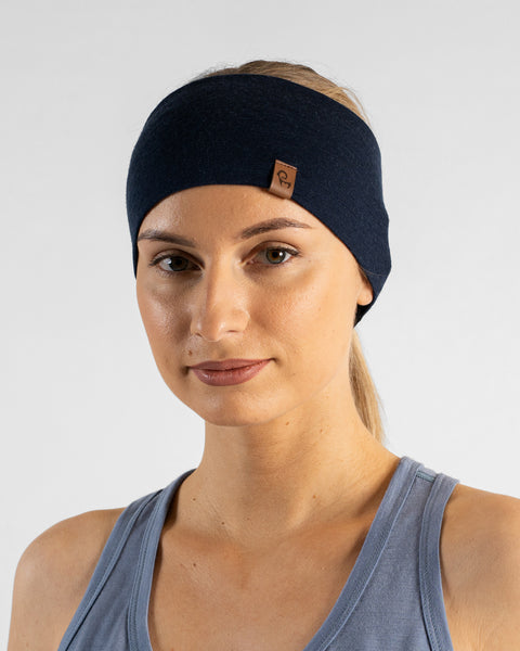 A studio portrait of a blonde woman looking directly at the camera. She is wearing a dark navy blue headband over her pulled-back hair, featuring a small brown leather logo patch. She is wearing a dusty blue sleeveless top against a plain light grey background.