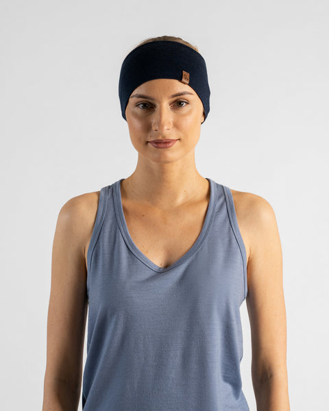 A studio portrait of a blonde woman facing forward and looking directly at the camera against a plain light grey background. She is wearing a dusty blue v-neck sleeveless top and a navy blue headband featuring a small brown logo tag.