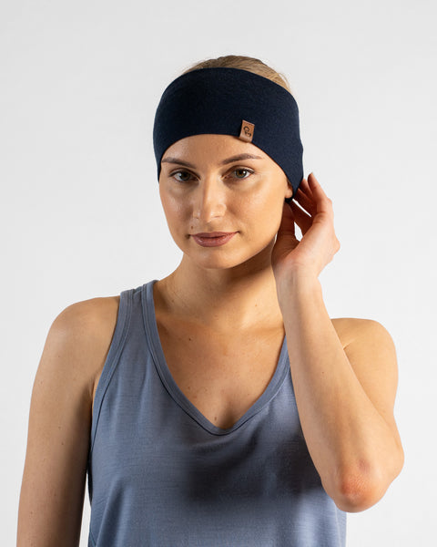 A studio portrait of a blonde woman facing forward and looking directly at the camera. She is wearing a dusty blue sleeveless top and a navy blue headband with a small brown logo tag, gently touching the edge of the headband near her ear with one hand against a plain light grey background.
