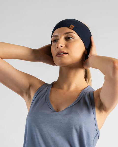 A studio portrait of a blonde woman looking off to the left and slightly upward against a plain light grey background. She is wearing a dusty blue sleeveless top and a navy blue headband, with both hands reaching up behind her head to adjust the fabric.