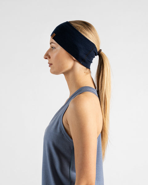 A side profile studio portrait of a blonde woman facing left against a plain light grey background. She is wearing a dusty blue sleeveless top and a navy blue headband over a ponytail, with a small brown logo tag visible on the side.