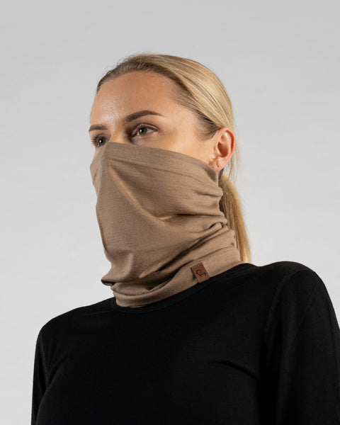 A studio portrait of a blonde woman shown in a three-quarter profile looking off to the left against a light grey background. She is wearing a black long-sleeved shirt and a beige neck gaiter pulled up over her nose and mouth, with a small brown logo tag visible at the bottom edge.