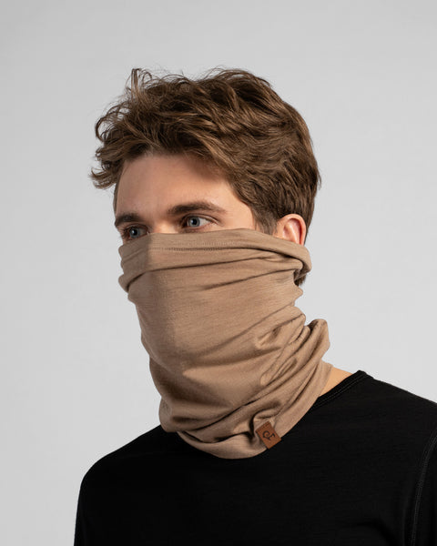 A studio portrait of a man looking off to the left against a light grey background. He is wearing a black shirt and a beige neck gaiter pulled up to cover his mouth and nose.