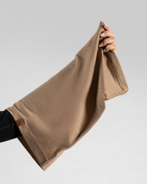 A close-up studio shot showing a person's hand holding up a beige tubular neck gaiter to display its full length and stretch, featuring a small brown logo tag at the bottom hem, against a plain light grey background.
