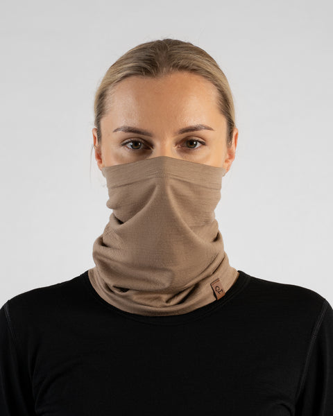 A studio portrait of a blonde woman facing forward and looking directly at the camera against a plain light grey background. She is wearing a black long-sleeved shirt and a beige neck gaiter pulled up to cover her nose and lower face, featuring a small brown logo tag at the collarbone.