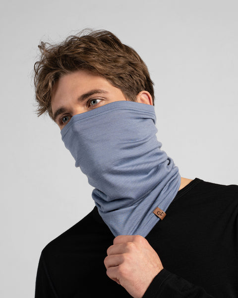 A studio portrait of a man looking thoughtfully off to the left side of the frame. He is wearing a dusty blue neck gaiter pulled up over his nose, covering his lower face. His right hand is holding the bottom edge of the gaiter, which features a small brown leather logo patch. He is wearing a solid black long-sleeved top against a plain light grey background.