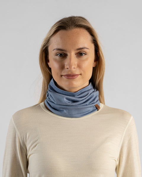 A studio portrait of a blonde woman facing forward and looking directly at the camera against a plain light grey background. She is wearing a light beige long-sleeved shirt and a dusty blue neck gaiter bunched around her neck, featuring a small brown logo tag.
