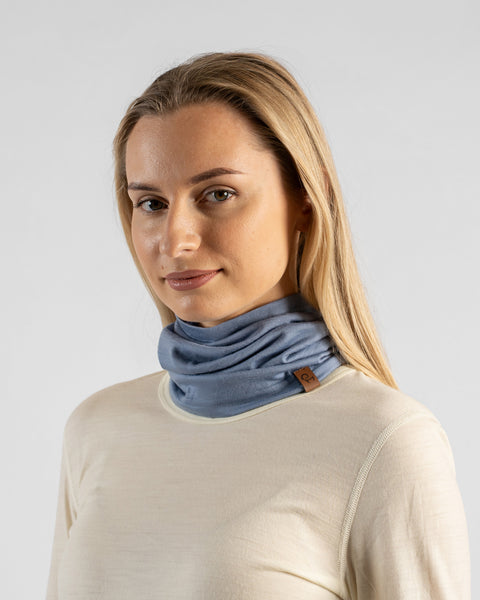 A studio portrait of a blonde woman shown in a three-quarter profile facing left, looking directly at the camera against a light grey background. She is wearing a light beige long-sleeved shirt and a dusty blue neck gaiter scrunched around her neck, featuring a small brown logo tag.