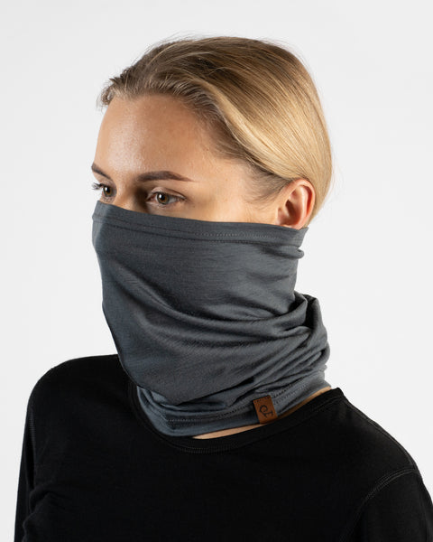 A studio portrait of a blonde woman facing left in a three-quarter profile against a plain light grey background. She is wearing a black long-sleeved shirt and a perfect gray neck gaiter pulled up to cover her nose and mouth, featuring a small brown logo tag at the base.