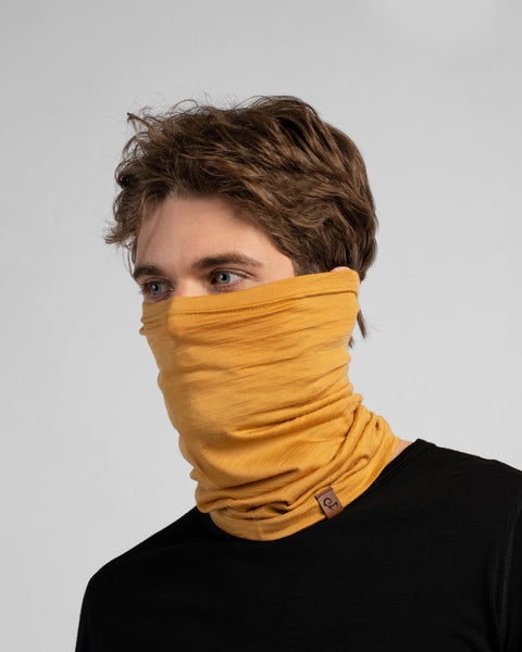 A studio portrait of a man looking thoughtfully off to the left side of the frame. He is wearing a mustard yellow neck gaiter pulled up over his nose, covering his lower face. The gaiter has a small brown leather logo patch on the bottom edge. He is wearing a solid black long-sleeved top against a plain light grey background.