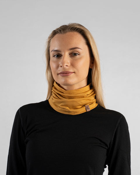 A studio portrait of a blonde woman facing forward and looking directly at the camera against a plain light grey background. She is wearing a black long-sleeved shirt and a mustard yellow neck gaiter bunched loosely around her neck, featuring a small brown logo tag.