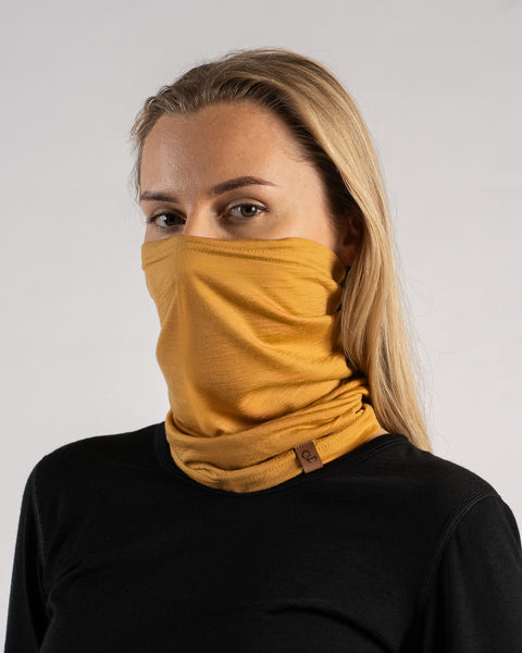 A studio portrait of a blonde woman looking directly at the camera. She is wearing a mustard yellow neck gaiter pulled up to cover her nose and lower face, along with a solid black long-sleeved top. A small brown leather logo patch is visible on the bottom edge of the gaiter. The background is a plain light grey.