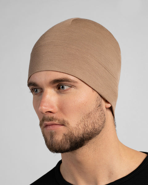 A close-up studio portrait of a man looking thoughtfully off to the left side of the frame. He is wearing a fitted, light beige skull cap and a solid black crew-neck top. The background is a plain light grey.