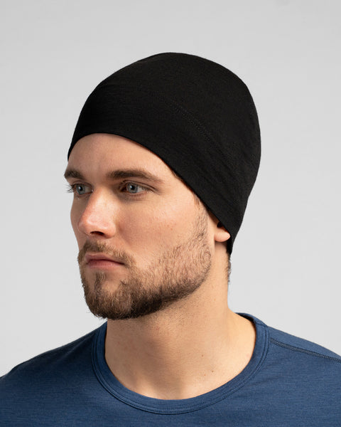 A close-up studio portrait of a man looking thoughtfully off to the left side of the frame. He is wearing a fitted, solid black skull cap and a dark blue crew-neck top. The background is a plain light grey.