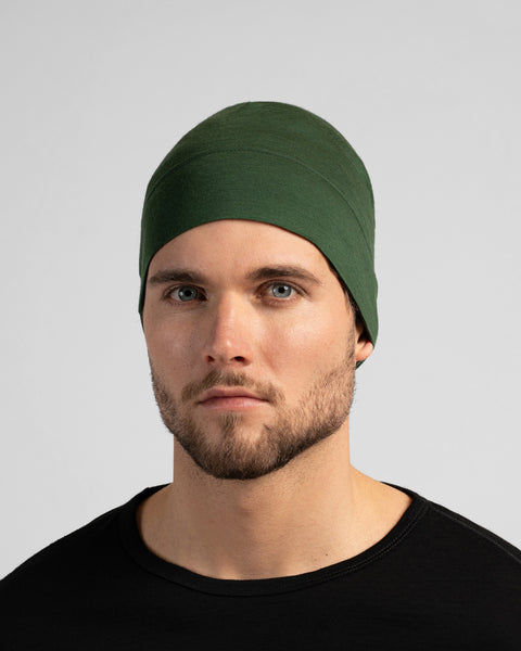 A close-up studio portrait of a man looking directly at the camera. He is wearing a fitted, dark green skull cap and a solid black crew-neck top. The background is a plain light grey.