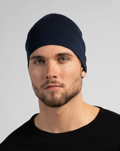 A close-up studio portrait of a man looking directly at the camera. He is wearing a fitted, dark navy blue skull cap and a solid black crew-neck top. The background is a plain light grey.