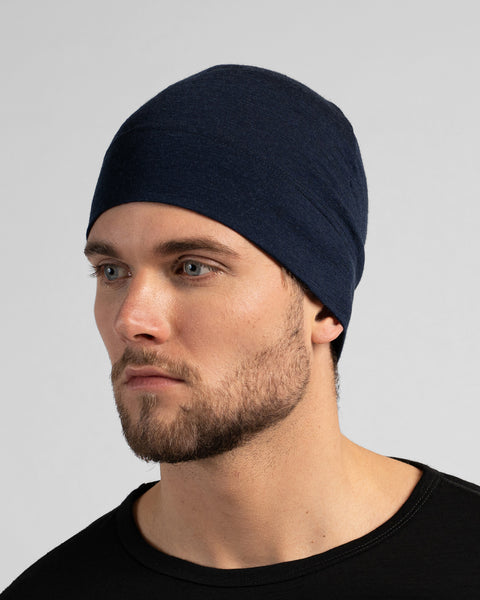 A close-up studio portrait of a man looking thoughtfully off to the left side of the frame. He is wearing a fitted, dark navy blue skull cap and a solid black crew-neck top. The background is a plain light grey.