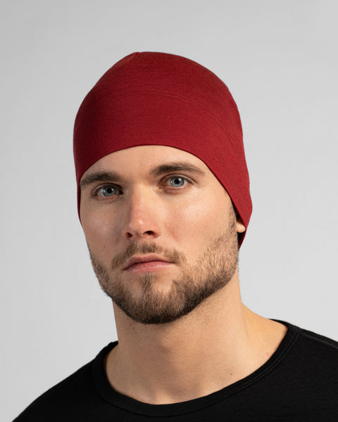 A close-up studio portrait of a man looking directly at the camera. He is wearing a fitted, dark red (royal cherry) skull cap and a solid black crew-neck top. The background is a plain light grey.