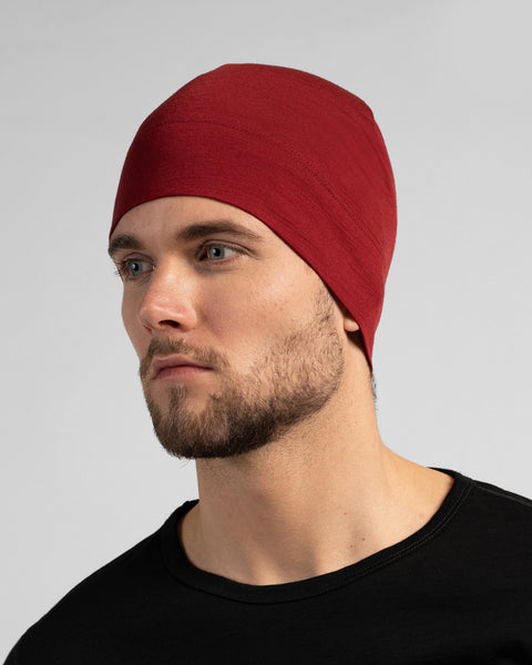 A close-up studio portrait of a man looking thoughtfully off to the left side of the frame. He is wearing a fitted, dark red (royal cherry) skull cap and a solid black crew-neck top. The background is a plain light grey.