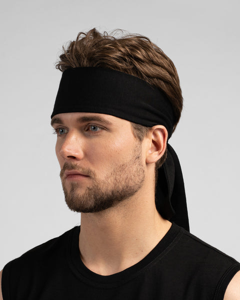 A studio portrait of a man looking thoughtfully off to the left side of the frame. He is wearing a solid black tie-back headband, with the tied ends hanging down behind his neck. He is wearing a black sleeveless top against a solid light grey background.