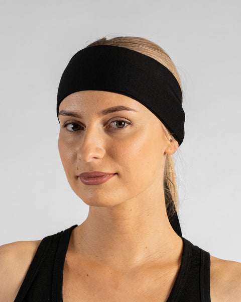 A studio portrait of a woman looking directly at the camera with a gentle expression. She is wearing a solid black tie-back headband over her blonde ponytail and a black tank top. The background is a solid light grey.