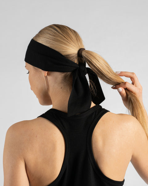 A studio portrait of a blonde woman facing away from the camera, showing her back and the back of her head against a plain light grey background. She is wearing a black sleeveless racerback top and a black tie headband tied over her ponytail, with one hand gently holding the end of her ponytail.