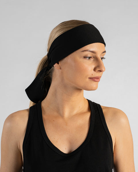 A studio portrait of a blonde woman shown in a three-quarter profile facing right, looking off-camera against a plain light grey background. She wears a black sleeveless top and a black tie headband with the tail ends hanging down behind her neck.