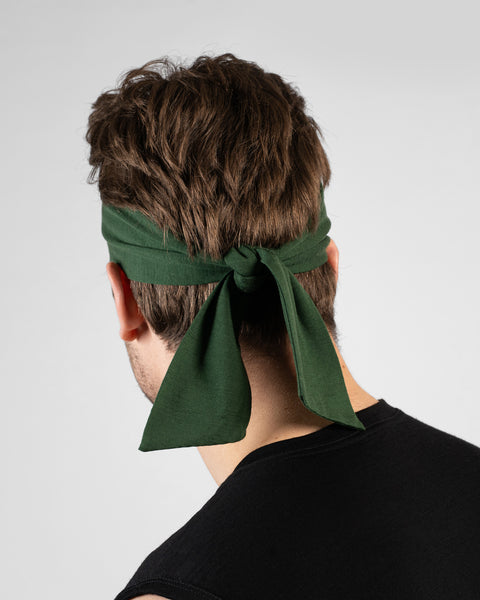 A rear-view studio portrait of a man's head and shoulders. He is shown from behind, wearing a dark green tie-back headband. The headband is tied in a knot at the back of his head with the ends hanging down. He is wearing a black sleeveless top against a solid light grey background.
