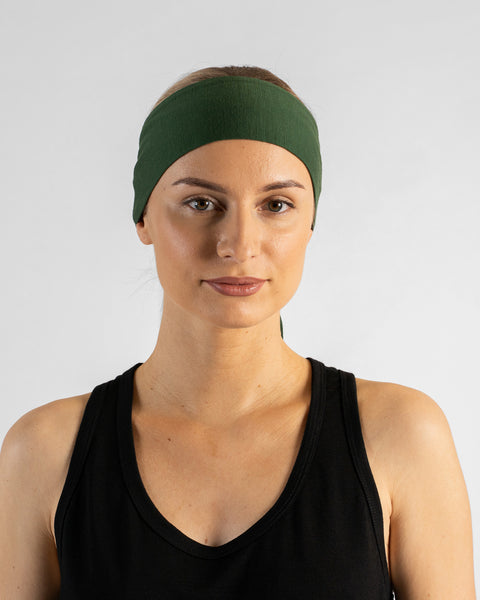 A studio portrait of a blonde woman facing forward and looking directly at the camera against a plain light grey background. She wears a black sleeveless top and a dark green tie headband with the tail ends hanging down behind her neck.