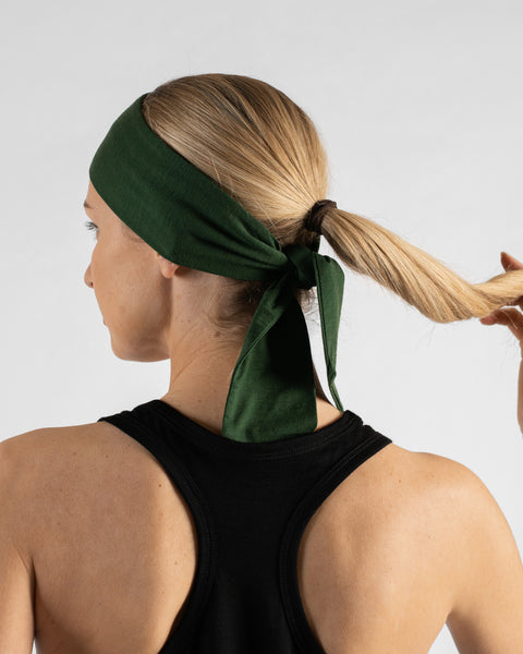 A studio portrait showing the back of a blonde woman against a plain light grey background. She is wearing a black sleeveless racerback top and a dark green tie headband tied over her ponytail, with one hand gently holding the end of her ponytail.