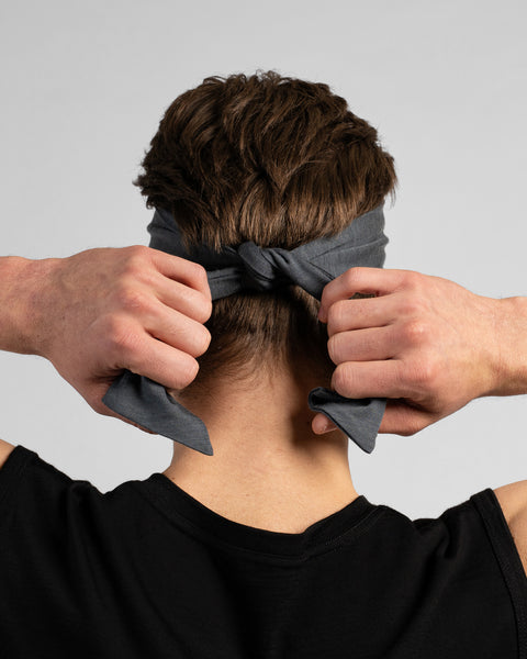 A rear-view studio portrait of a man's head and shoulders. He is shown from behind, using both hands to tie a medium grey headband at the back of his head. He is wearing a black sleeveless top against a solid light grey background.