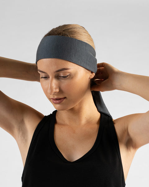 A studio portrait of a blonde woman looking downward and to the left, with both hands raised behind her head to tie a perfect gray headband. She is wearing a black sleeveless v-neck top against a plain light grey background.