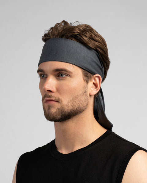 A studio portrait of a man looking thoughtfully off to the left side of the frame. He is wearing a medium grey tie-back headband, with the tied ends hanging down behind his neck. He is wearing a black sleeveless top against a solid light grey background.