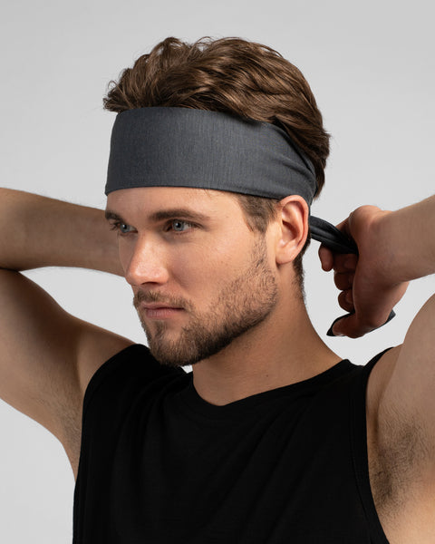 A man with wavy brown hair looks to the left while tying a grey headband behind his head. He is wearing a black tank top against a plain light grey background.