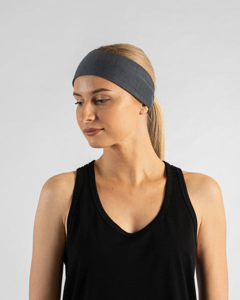 A studio portrait of a blonde woman looking downward and to the left against a plain light grey background. She is wearing a black sleeveless v-neck top and a perfect gray tie headband over a ponytail.