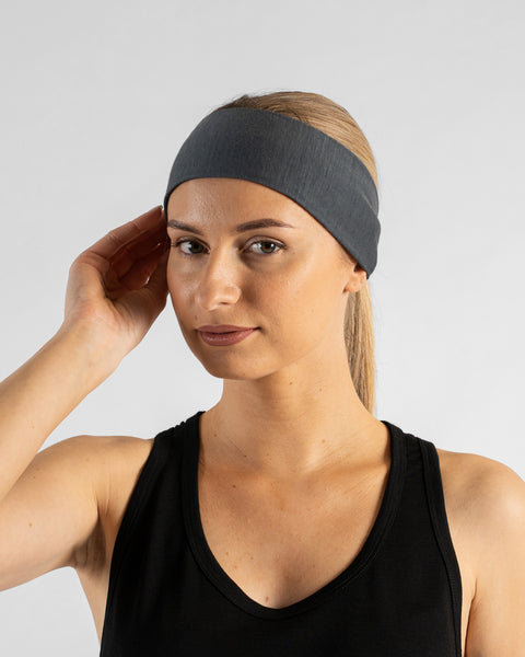 A studio portrait of a woman looking directly at the camera with a subtle smile. She is wearing a medium grey tie-back headband over her blonde hair and a black tank top. Her right hand is gently resting on the side of the headband. The background is a solid light grey.