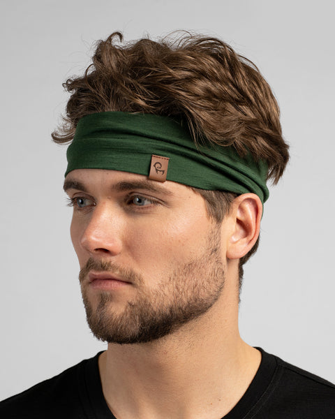 A close-up studio portrait of a man looking to the left against a plain light grey background. He has wavy brown hair held back by a dark green headband and is wearing a black shirt.