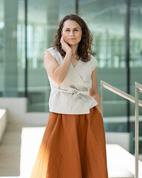 A woman with brown hair, wearing a light beige wrap top and a long rust-brown skirt, poses indoors in front of large windows.