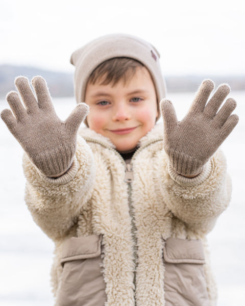 A smiling young boy holding both hands open towards the camera, showcasing light beige knitted gloves. He wears a matching light beige beanie and a fuzzy cream-colored fleece jacket. The background is a softly blurred outdoor winter scene.